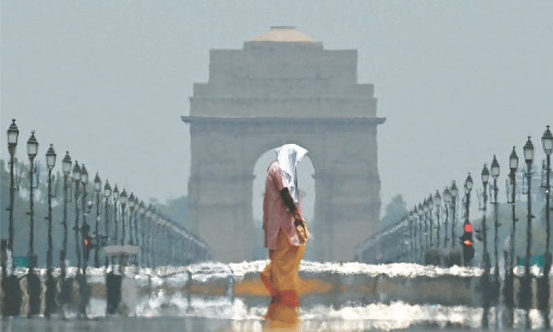 A man crossing a rain-soaked road with a cloth on his head during Delhi monsoon, India Gate in the background — best time to visit in Kerala.
